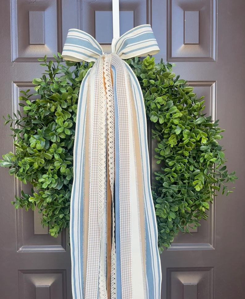 May include: A green boxwood wreath with a blue and white striped bow and cascading ribbons. The ribbons include lace, gingham, and solid colors of blue, beige, and white. The wreath hangs on a brown door.