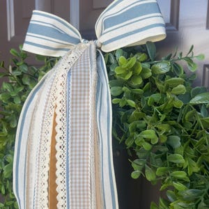 May include: A decorative wreath featuring a lush green base, adorned with a large bow. The bow is made of striped blue and white fabric, with coordinating ribbons in beige, cream, and lace. This wreath is perfect for adding a touch of farmhouse charm to any home.
