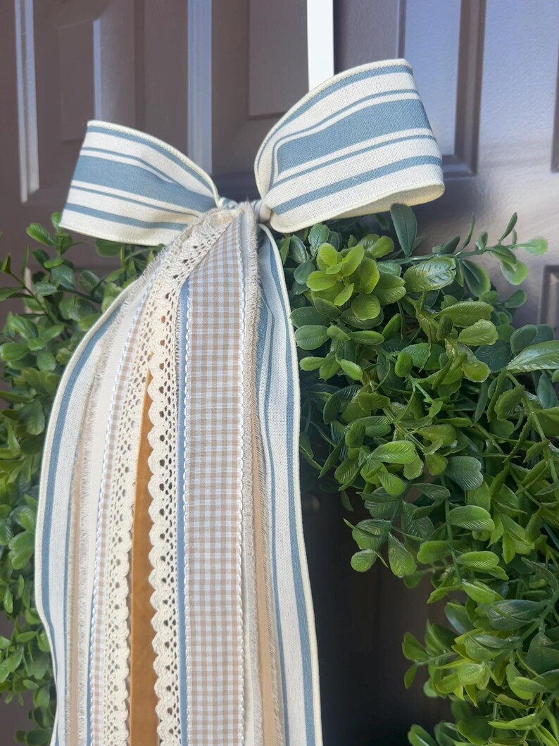 May include: A decorative wreath featuring a lush green base, adorned with a large bow. The bow is made of striped blue and white fabric, with coordinating ribbons in beige, cream, and lace. This wreath is perfect for adding a touch of farmhouse charm to any home.