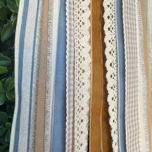 May include: A close-up of various ribbons and trims in a vertical arrangement. The ribbons include blue and white striped fabric, beige gingham, cream lace, and a mustard-colored velvet ribbon. The textures and colors create a layered, decorative effect.