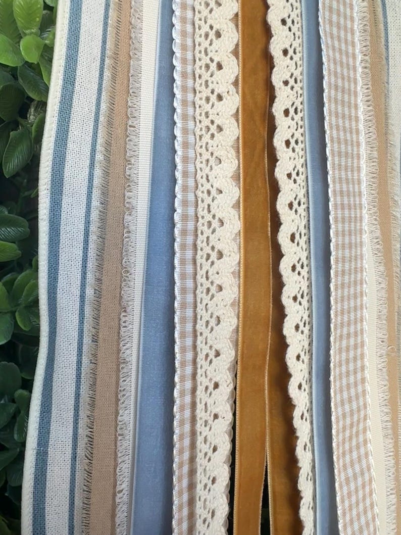 May include: A close-up of various ribbons and trims in a vertical arrangement. The ribbons include blue and white striped fabric, beige gingham, cream lace, and a mustard-colored velvet ribbon. The textures and colors create a layered, decorative effect.