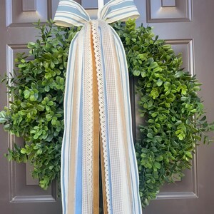 May include: A green wreath with a variety of ribbons and a bow. The ribbons are in shades of blue, tan, and white, with lace and striped patterns. The bow is blue and white striped. The wreath is hanging on a brown door.