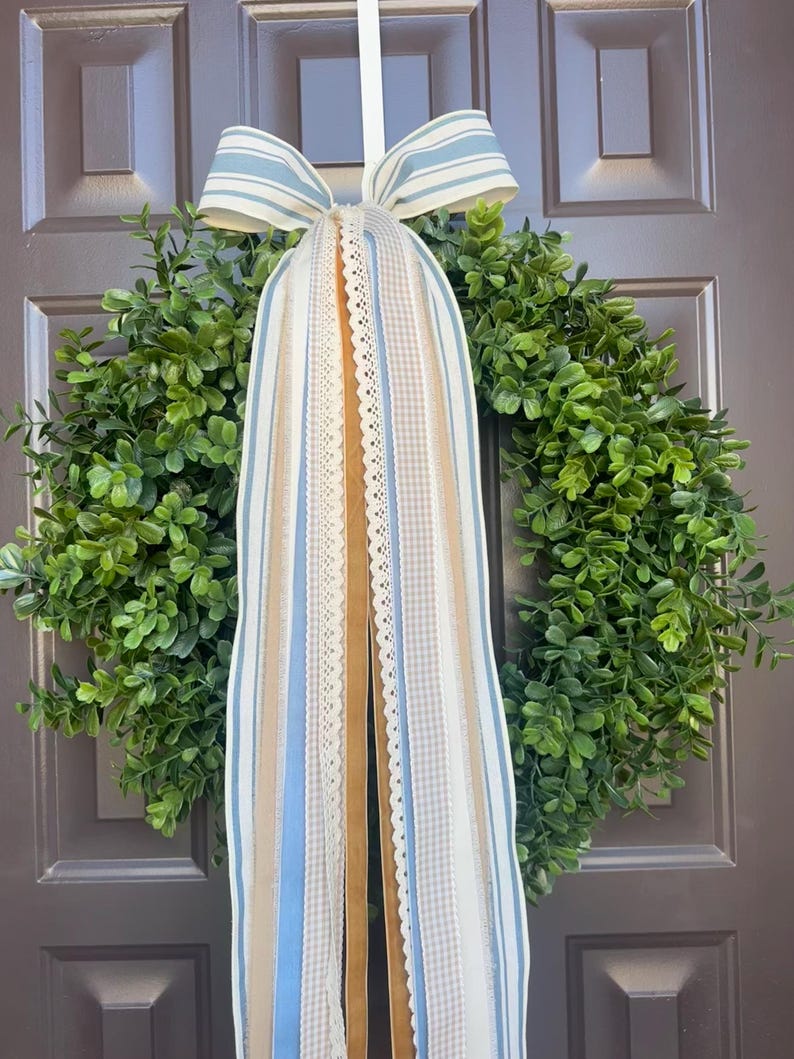May include: A green wreath with a variety of ribbons and a bow. The ribbons are in shades of blue, tan, and white, with lace and striped patterns. The bow is blue and white striped. The wreath is hanging on a brown door.