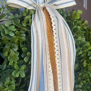 May include: A decorative wreath with a lush green base and a large bow. The bow features blue and white striped fabric, along with various ribbons in cream, tan, and gold. The ribbons include lace and gingham patterns.