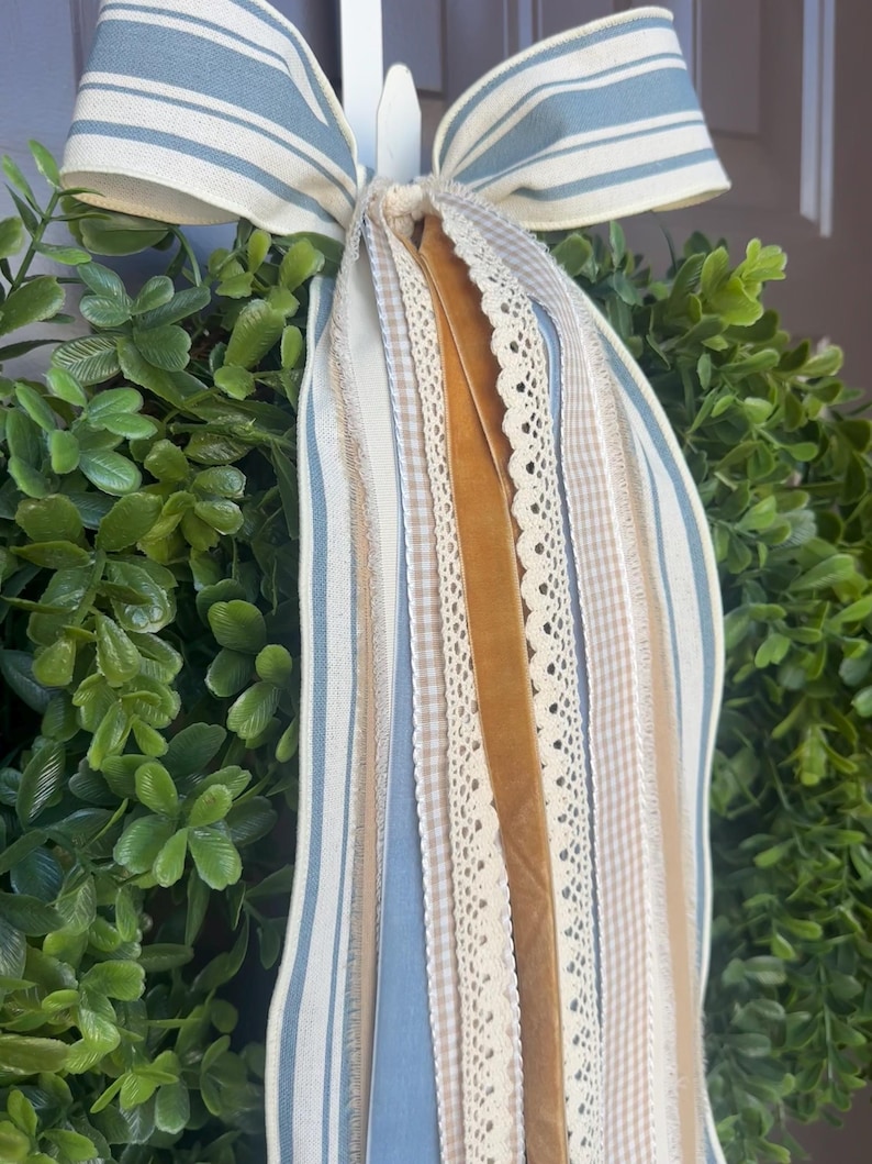 May include: A decorative wreath with a lush green base and a large bow. The bow features blue and white striped fabric, along with various ribbons in cream, tan, and gold. The ribbons include lace and gingham patterns.