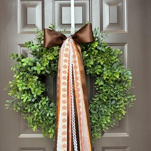 May include: A circular green boxwood wreath featuring a large brown satin bow at the top. Several long, hanging ribbons in various textures and patterns, including orange pumpkin prints, brown gingham, and white lace, drape down from the center.