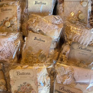 May include: Multiple clear plastic bags containing assorted cream and white buttons and lace fabric scraps. Each bag features a card with the word Buttons printed on it, some with floral designs, containing various sizes of round buttons.
