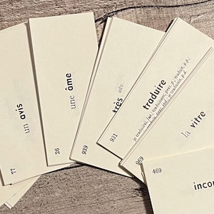 May include: A collection of cream-colored flashcards with French vocabulary words printed in black. The cards are fanned out on a wooden surface, with words like "le pied" and "traduire" visible.