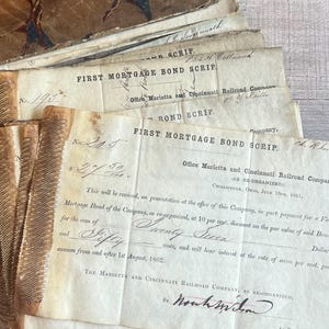 May include: A stack of aged paper documents, featuring the text "FIRST MORTGAGE BOND SCRIP" and handwritten notations. The papers have a vintage aesthetic, with visible wear and tear, and are likely from the Marietta and Cincinnati Railroad Company.