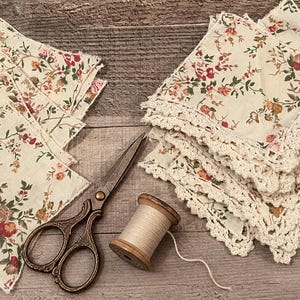 May include: A collection of square fabric napkins featuring a cream background with a floral pattern in red, orange, and green. One stack of napkins has a decorative cream crochet border. A pair of bronze-toned metal scissors and a wooden spool of thread.
