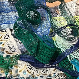 May include: A collection of various lace ribbons in shades of blue, green, and white. The ribbons have different patterns and textures, including floral and geometric designs. Some ribbons are solid colors, while others have a gradient effect.