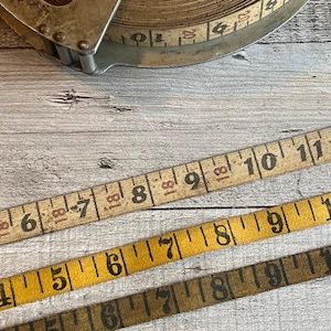May include: Close-up of vintage measuring tapes in various shades of tan and yellow. The tapes are marked with black numbers and inch increments. A metal tape measure reel is visible in the background.