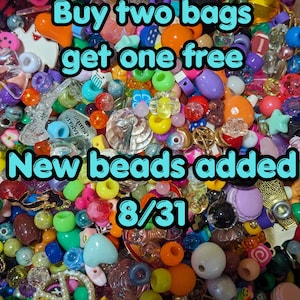 May include: A large collection of assorted colorful plastic and metal craft beads in various shapes including hearts, stars, and flowers, with text overlay reading Buy two bags get one free New beads added 8/31 in light blue letters.