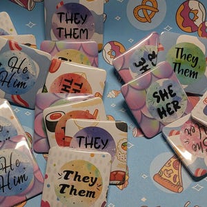 May include: A collection of square pronoun pins with watercolor backgrounds. The pins feature phrases like "He/Him", "She/Her", and "They/Them". The background is a light blue pattern with food illustrations.