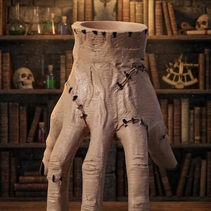 May include: A beige, hand-shaped ceramic vase with black fingernails and stitched details. The open top of the vase is lined with small holes. The background features a library with bookshelves, books, and skulls.