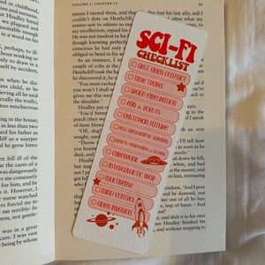 May include: A white bookmark with red text that reads "SCI-FI CHECKLIST." The checklist includes topics like "First Alien Contact" and "Alien Invasion." A rocket and planet graphic are at the bottom. The bookmark is on top of an open book.