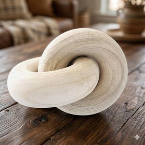 May include: A light-colored wooden decorative chain link sculpture. The interlocking rings have a smooth, rounded shape and a natural wood grain texture. The sculpture is displayed on a rustic wooden table.
