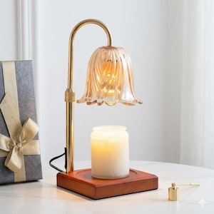 May include: A decorative candle warmer with a gold-colored metal frame and a flower-shaped glass shade. The warmer sits on a wooden base and includes a lit candle. A gift box with a gold ribbon is in the background.