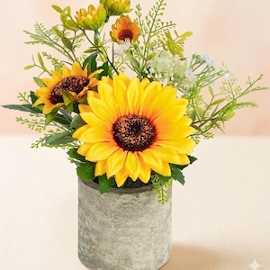 May include: A vibrant arrangement of sunflowers and greenery in a gray concrete-look pot. The sunflowers have bright yellow petals and dark brown centers. The arrangement includes other yellow and white flowers and green foliage.