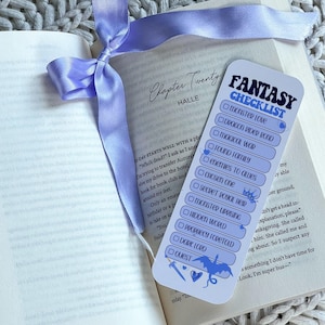 May include: A bookmark with a fantasy checklist design, featuring a blue dragon illustration and the text "Fantasy Checklist." The bookmark is placed on an open book with a light purple ribbon.