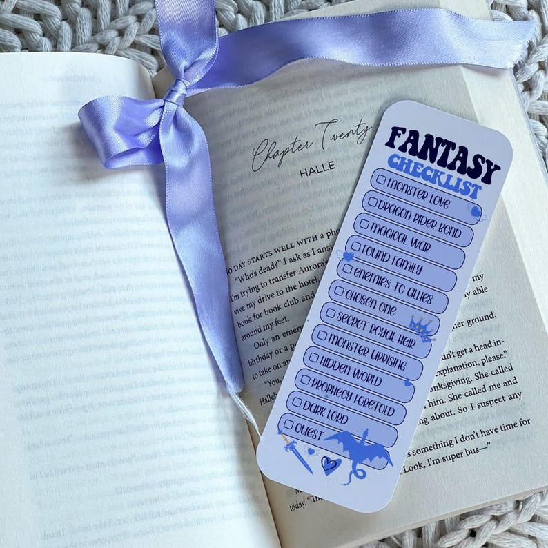 May include: A bookmark with a fantasy checklist design, featuring a blue dragon illustration and the text "Fantasy Checklist." The bookmark is placed on an open book with a light purple ribbon.
