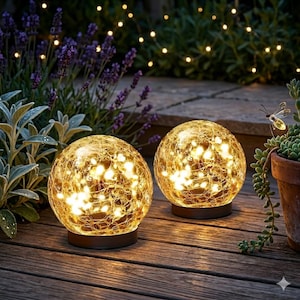 May include: Two illuminated glass globe lights with a crackled texture, resting on brown bases. The globes are filled with warm white lights, creating a cozy ambiance. Set on a wooden deck, surrounded by greenery and string lights.