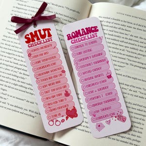 May include: Two pink and white bookmarks with text. One says "SMUT CHECKLIST" with phrases like "Forced Proximity." The other says "ROMANCE CHECKLIST" with phrases like "Friends to Lovers." A maroon ribbon is attached to the top.