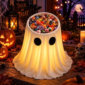 May include: A white, ghost-shaped candy bowl with two black circular eyes, illuminated from within, filled with a variety of colorful wrapped candies. The bowl features a draped fabric appearance and serves as a container for treats.