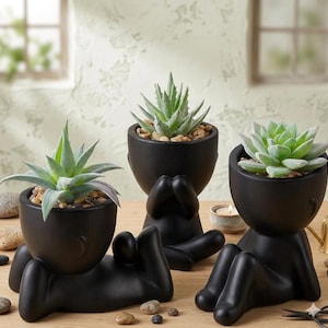 May include: Three black ceramic planters shaped like figures, each holding a succulent plant. The planters have a minimalist design with a smooth, matte finish. The succulents are green and surrounded by small pebbles.