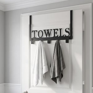 May include: Black metal over-the-door towel rack with the word "TOWELS" cut out. Two towels hang from the rack: one white and one gray. The rack is mounted on a white door in a bathroom setting.
