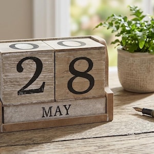 May include: Wooden block calendar displaying "28 MAY" with large black numbers and text. The calendar sits on a wooden table next to a small potted plant and a pen. The calendar is a light brown color.