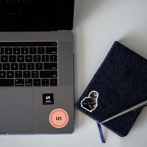 May include: A laptop, a navy blue journal, and a pen are on a white surface. Two stickers are on the laptop, one black with white text and the other a peach circle with the letters "un". A sticker of a person in a racing suit is on the journal.