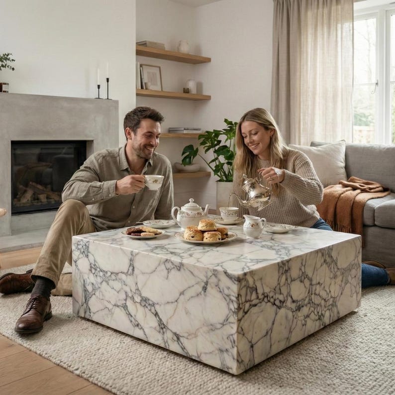 May include: A marble coffee table with a white and gray veined pattern is the focal point of a living room scene. A couple is seated around the table, enjoying tea and pastries. The room features a fireplace, neutral-toned rug, and natural light.