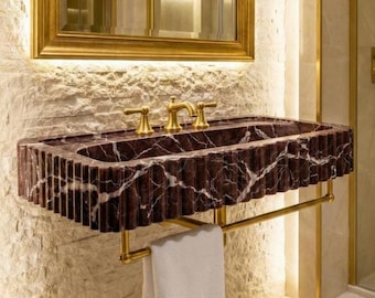 May include: A luxurious bathroom sink made of dark red marble with white veining. The sink has a fluted edge and gold-colored fixtures, including faucets and a towel rack. A gold-framed mirror hangs above the sink, and a white towel is on the rack.