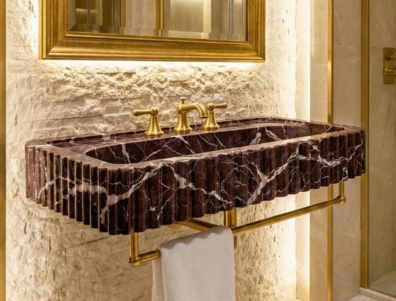 May include: A stylish bathroom sink crafted from dark red marble with white veining. The sink features a fluted edge and gold-coloured fixtures, including taps and a towel rail. A gold-framed mirror is above the sink, and a white towel hangs on the rail.