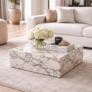 May include: A square marble coffee table with a white and gray veined pattern. A glass bowl of white flowers, books, a black candle, and decorative beads sit on top. The table is in a living room setting.