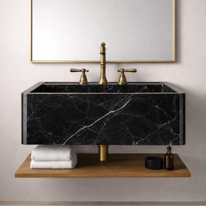 Op de afbeelding: Een rechthoekige wastafel van zwart marmer met witte adering, met een gouden kraan en handgrepen. De wastafel is gemonteerd boven een houten plank met opgevouwen witte handdoeken. Een goudomrande spiegel hangt erboven.