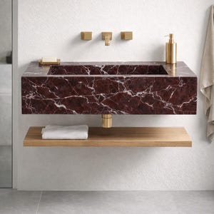 May include: A contemporary bathroom sink crafted from red marble with white veining. The rectangular sink is wall-mounted, featuring gold fixtures such as a tap and soap dispenser. A wooden shelf below holds a folded white towel.