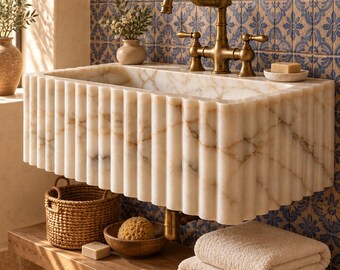 Puede incluir: Un lavabo rectangular montado en la pared, hecho de mármol blanco con un diseño exterior acanalado. El lavabo cuenta con un grifo de latón de dos manijas y se sitúa sobre un estante de madera con toallas blancas y una cesta tejida.