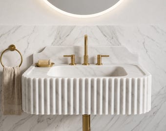May include: A white marble wall-mounted sink with a fluted front design, featuring a gold faucet, two gold handles, and a gold drain pipe. A gold soap dish with a bar of soap sits on the deck, and a beige towel hangs on a gold ring.