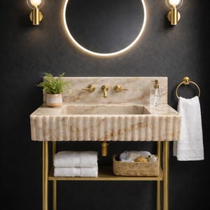 May include: A marble bathroom sink with gold fixtures, a round mirror, and sconces. The sink has a ribbed front and sits on a gold metal stand with a shelf holding towels and a basket. A white towel hangs on a gold ring.