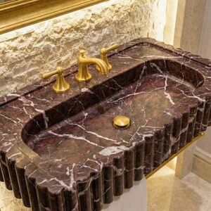 May include: A rectangular bathroom sink in dark red marble with white veining. The sink has a fluted edge and gold-coloured taps. The sink also features a gold-coloured drain stopper.