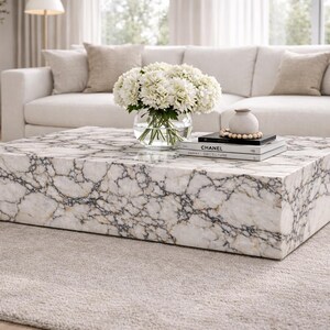 May include: A square marble coffee table with a white and gray veined pattern. A glass vase of white flowers, a small vase, and books are on the table. The table sits on a light gray rug in front of a white sofa.