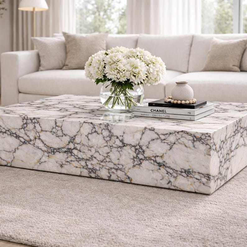 May include: A square marble coffee table with a white and gray veined pattern. A glass vase of white flowers, a small vase, and books are on the table. The table sits on a light gray rug in front of a white sofa.
