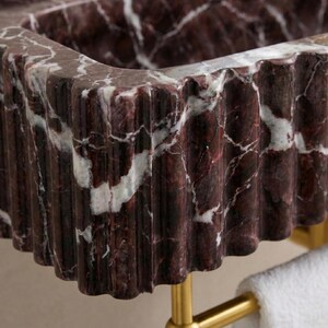 May include: Close-up of a burgundy marble sink with white veining. The sink has a fluted, wavy edge and is mounted on a gold-coloured metal frame. A white towel hangs below.