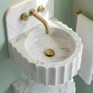 May include: A white marble sink with a scalloped edge and brass fixtures. The sink has a round basin and a curved tap. The fixtures include two knobs and a drain cover. A white towel hangs on a brass towel rail.
