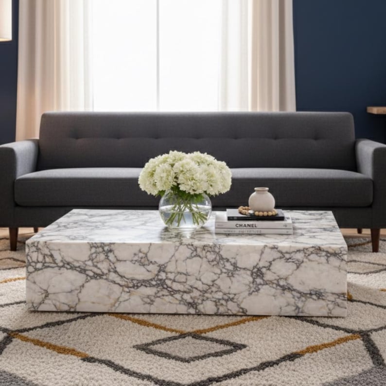 May include: A rectangular marble coffee table with gray veining, holding a glass vase of white flowers, a small white vase, and a book titled "CHANEL". A gray sofa sits in the background, with a patterned rug below.