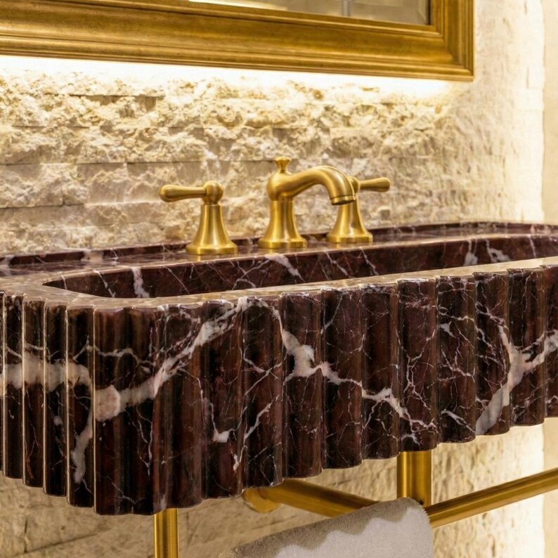 May include: A stylish bathroom sink crafted from dark red marble with white veining. The sink features a fluted design and is complemented by gold-toned taps and a towel rail. The background includes a textured stone wall and a gold-framed mirror.