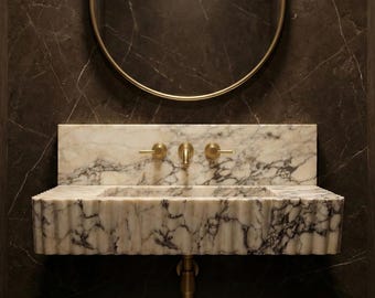 Puede incluir: Un lavabo de pared de mármol blanco con un diseño frontal acanalado y vetas gris oscuro. El lavabo cuenta con un grifo central de latón y dos manijas a juego, situado debajo de un espejo redondo con un marco fino de tono dorado.