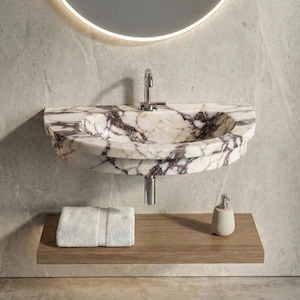 Puede incluir: Un lavabo de mármol con un patrón veteado blanco y morado. El lavabo está montado en una pared sobre una estantería de madera. Una toalla blanca y un dispensador de jabón están en la estantería. Un grifo cromado está instalado encima del lavabo.